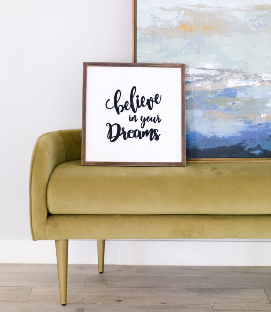 olive-colored velvet bench with abstract painting and framed words "believe in your dreams" sitting on it