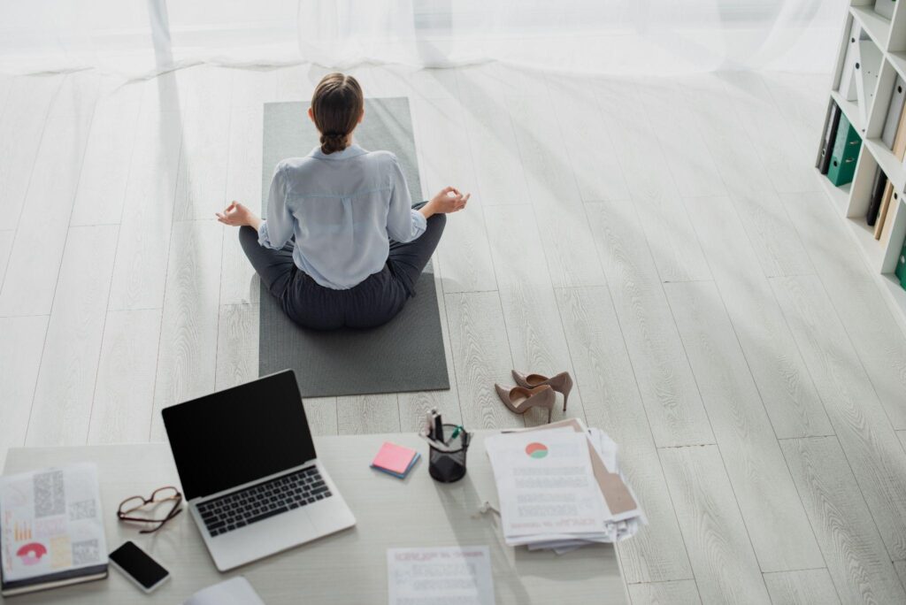 woman sitting on a yoga mat meditating. Desk with laptop, papers, office supplies in the foreground