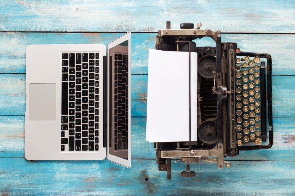 overhead view of laptop computer on left and antique manual typewriter sitting back to back on blue wood plank table