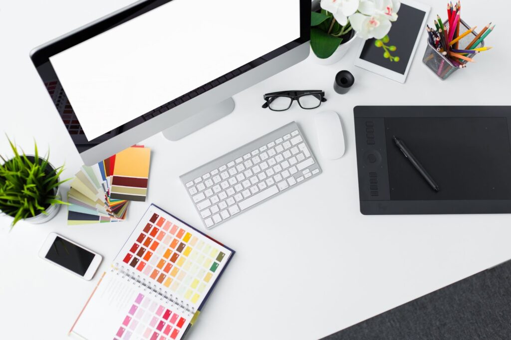 image containing items used in digital graphic design: tablet with stylus, computer monitor, keyboard, mouse, color swatches, colored pencils, cell phone; also included - 2 potted plants, eyeglasses